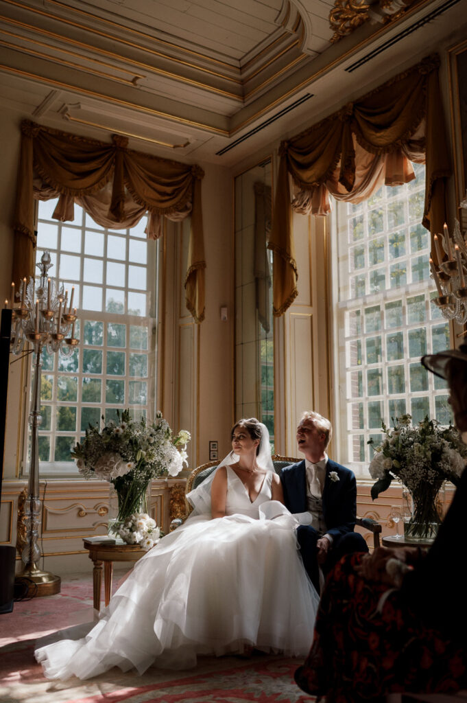 Bride and groom seated on a bench together inside an elegant wedding venue, with tall windows in the background
