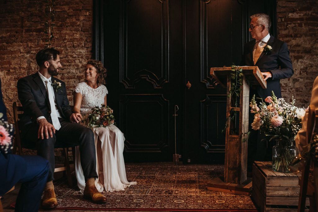binnenceremonie bruidspaar kijkt elkaar lachend aan terwijl ze worden toegesproken