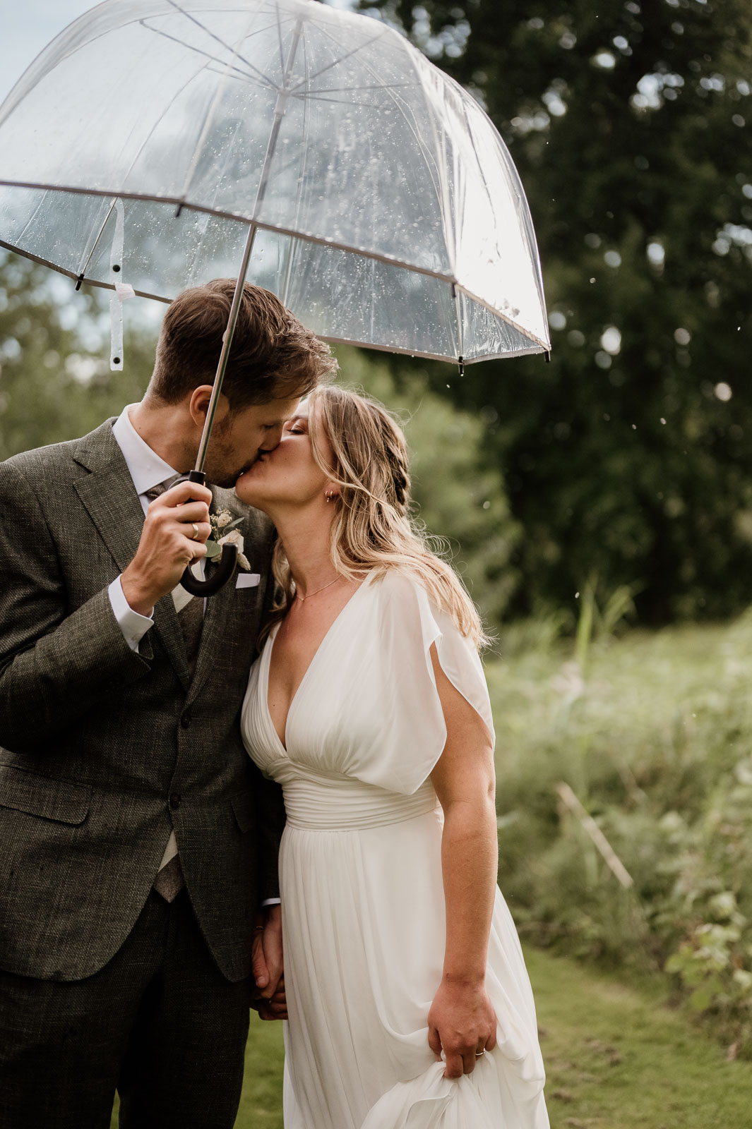 Couple in wedding attire kissing in the rain, the groom holding a white umbrella