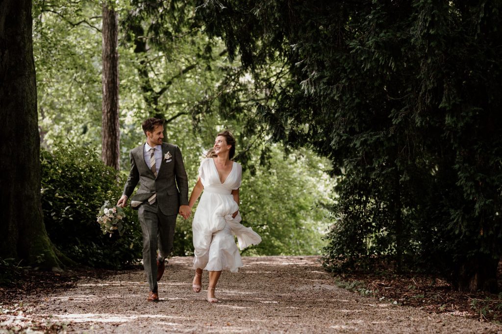 Couple in wedding attire running down a forest path, holding hands and smiling at each other