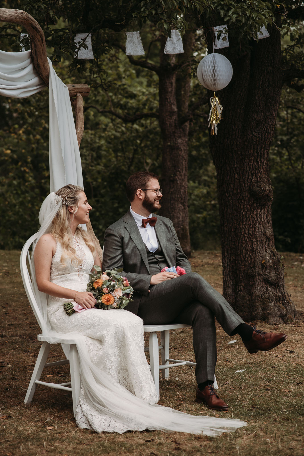 Wedding couple sitting in white chairs underneath a tree, looking to the side and smiling happily