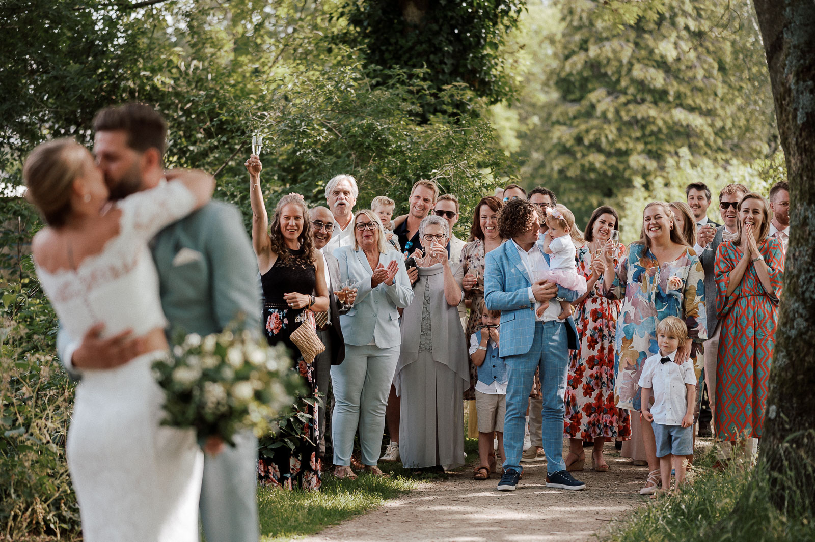 Couple in wedding attire kissing while wedding party cheers in the background