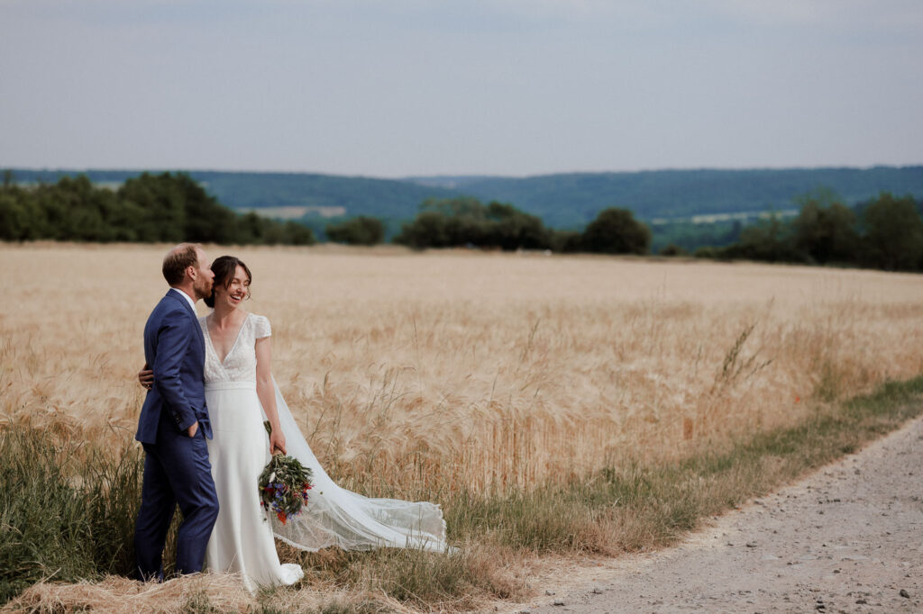 Wedding couple holding each other, the groom kissing the bride while she laughs, a field in the background