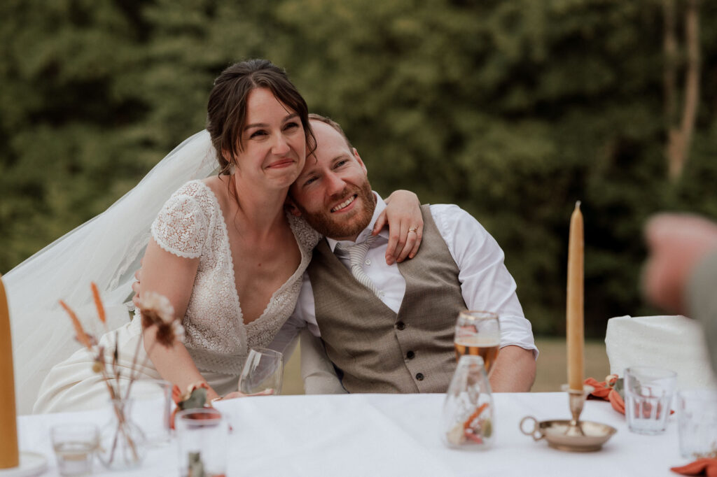 Wedding couple seated together at a dinner table, smiling while holding each other, with greenery in the background