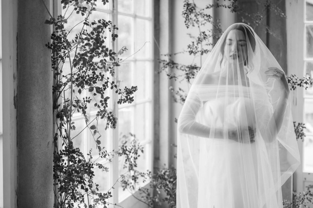 Bride wearing a long, white veil, her eyes closed, a tall window in the background
