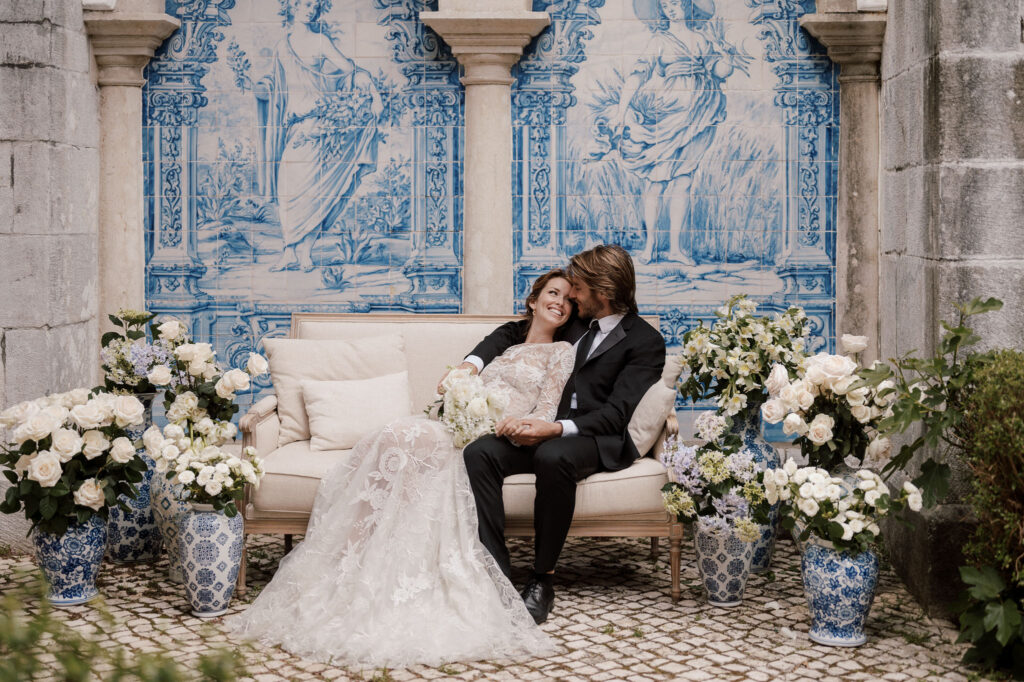 Wedding couple sitting together on a couch and smiling at each other, surrounded by Portuguese tiles and white flowers