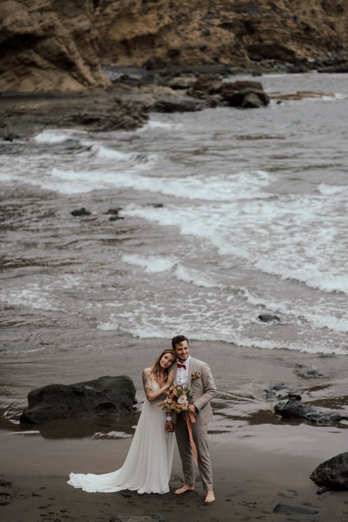wedding and elopement photography tenerife, Bride and groom embracing on a rocky beach with black sand and waves crashing behind them