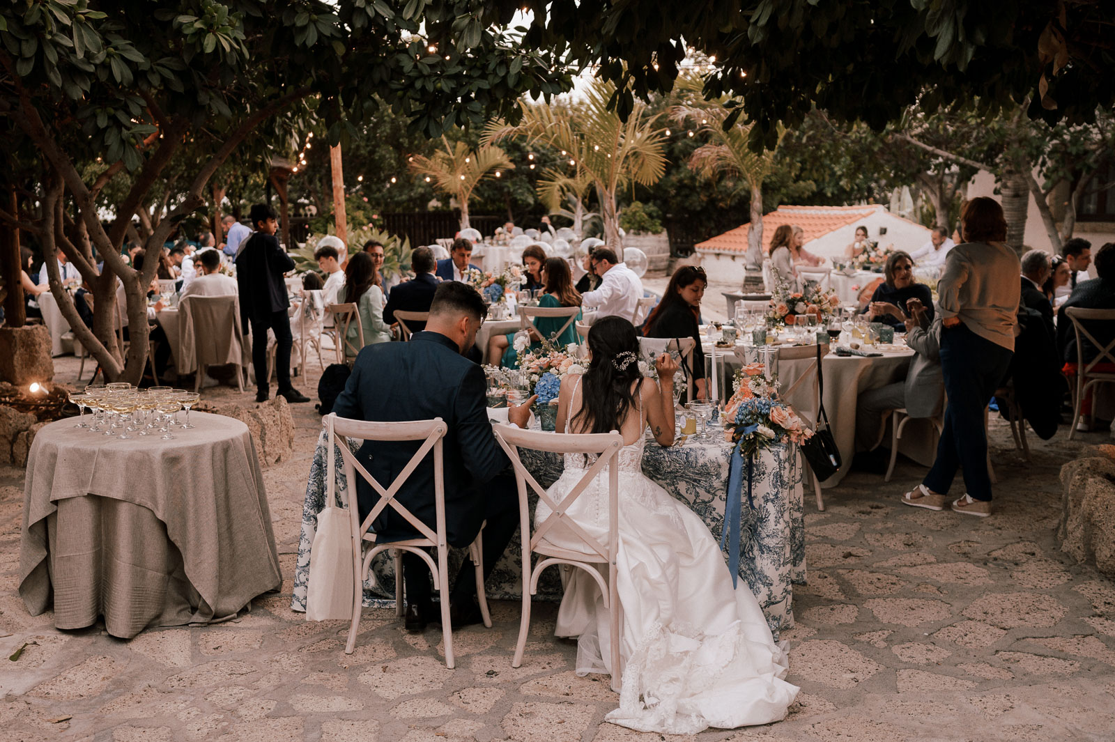 wedding and elopement photography, Outdoor wedding reception under the trees with the bride and groom seated at a flower-decorated table, surrounded by guests enjoying dinner and string lights
