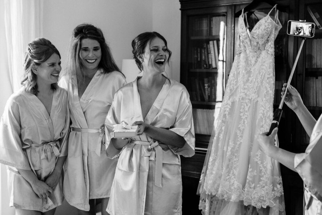 Bridesmaids in robes laughing together while taking a selfie with the wedding dress hanging nearby
