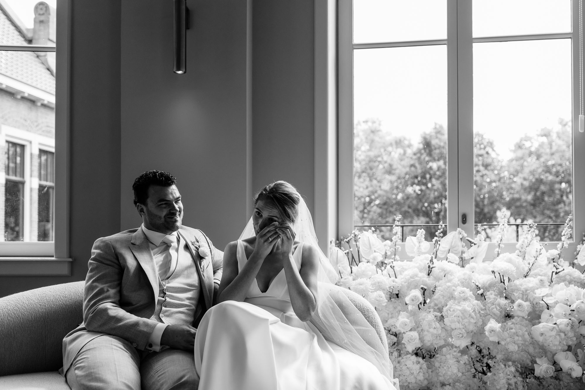 Bride wiping away tears while sitting beside the groom on a sofa, surrounded by elegant floral arrangements, wedding photography